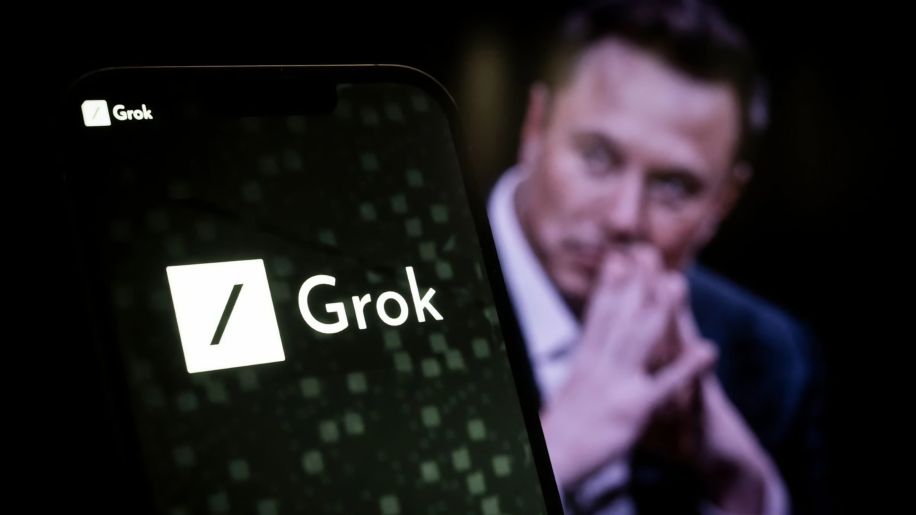 Ein Smartphone zeigt das Logo und den Schriftzug „Grok“ auf dem Bildschirm. Im unscharfen Hintergrund ist ein Mann mit gefalteten Händen zu sehen.