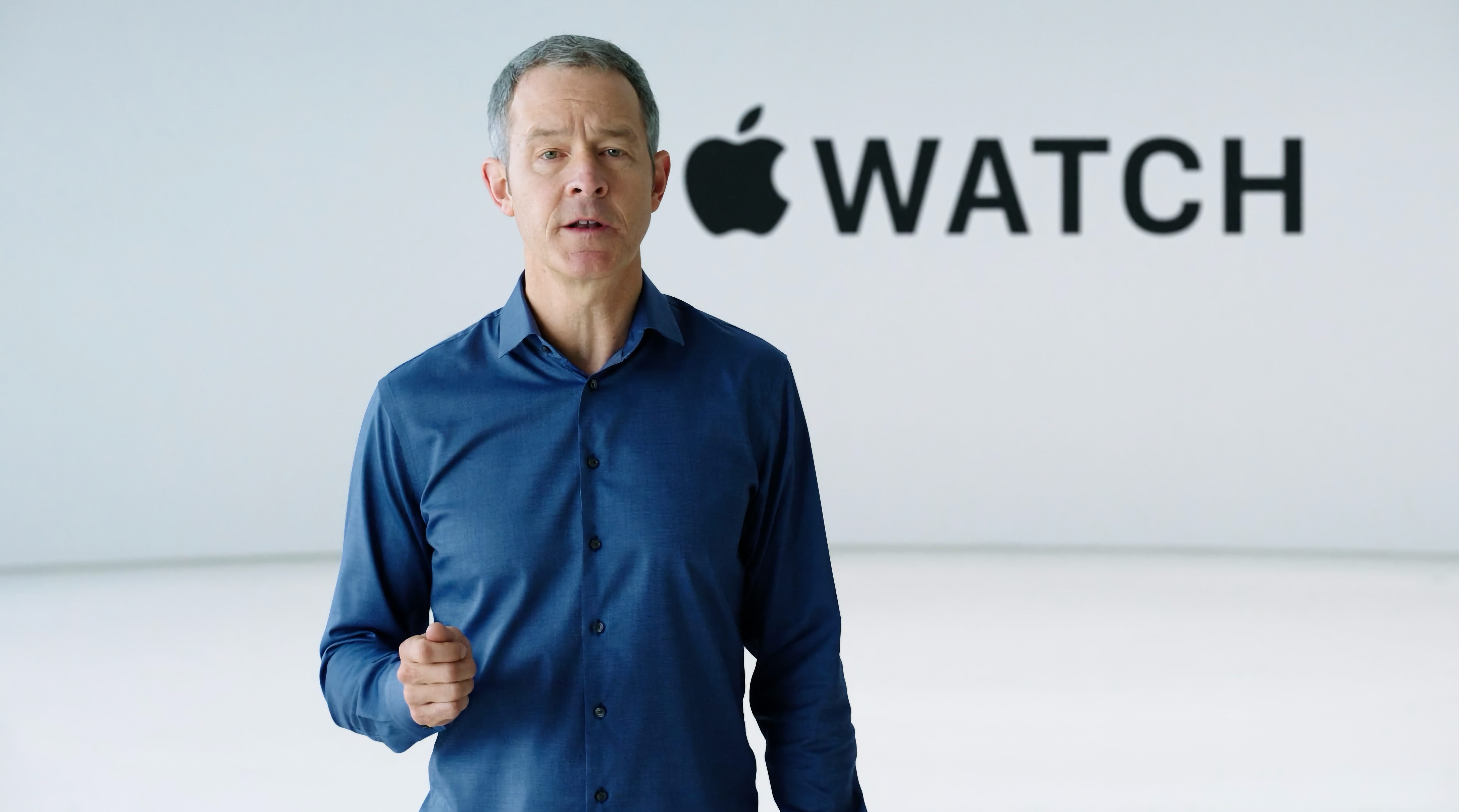 Ein Mann mit grauen Haaren trägt ein blaues Hemd und steht vor einer weißen Wand mit dem schwarzen Apple-Logo und dem Schriftzug „WATCH“.