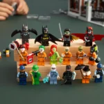 Mehrere LEGO-Figuren aus dem Batman-Universum, darunter Batman, Robin, Batgirl, Joker, Poison Ivy, Riddler und weitere, auf einem dreistufigen Holzpodest arrangiert. Im Hintergrund sind Hände, ein LEGO-Gebäude und ein Polizeiauto unscharf zu sehen.