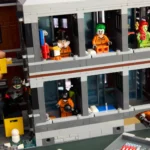 LEGO-Gebäude mit mehreren Minifiguren, darunter Joker mit grünen Haaren, Poison Ivy mit roten Haaren, ein Mann mit Zylinderhut und weitere Figuren in orangen Overalls. Eine Hand hält ein gelbes LEGO-Teil an der linken Seite des Gebäudes.