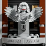 Eine graue Engelsfigur aus LEGO mit ausgebreiteten Flügeln, gesperrten Händen und einem Schloss an der Vorderseite. Die Figur hat ein Gesicht mit wirrem, langem Haar und einem gruseligen, verzerrten Gesichtsausdruck. Darunter steht der Schriftzug „ARKHAM ASYLUM“.