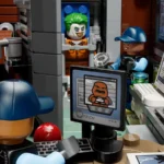 Zwei Lego-Figuren in blauen Kappen und Uniformen überwachen einen Gefängnisinsassen mit grünen Haaren und oranger Kleidung, der in einer Zelle steht. Auf einem Monitor ist ein weiteres Lego-Gesicht zu sehen, daneben ein Computer mit der Anzeige „OPEN ALL CELL DOORS?“. Im Hintergrund sind Gefängniselemente wie eine Tasse, ein Telefon und eine Messlatte an der Wand zu erkennen.