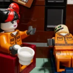 Zwei LEGO-Figuren in orangener Kleidung, eine sitzt in einem roten Stuhl mit einer weißen Tasse in der Hand, die andere liegt auf einer blauen Liege. Die sitzende Figur hat schwarz-rote Haare und lächelt, die liegende Figur hat ein bemaltes Gesicht mit grünen Augen. Hintergrund mit braunen und schwarzen Fliesen.