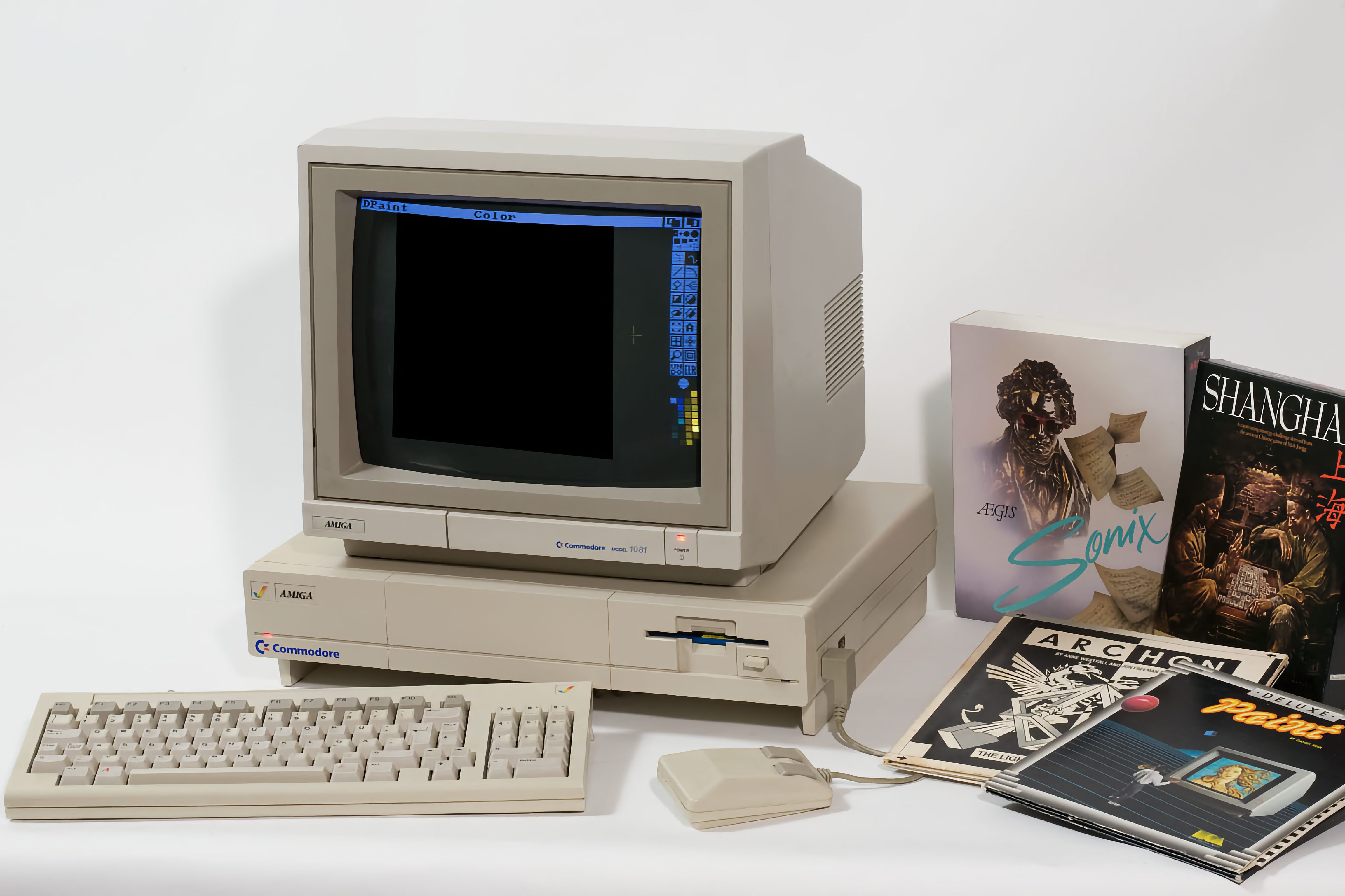 Commodore Amiga 1081 Monitor auf einem beigefarbenen Commodore Amiga Computer mit Tastatur und Maus, daneben mehrere Spiele- und Softwareverpackungen und Handbücher. Der Monitor zeigt ein geöffnetes Malprogramm namens "DPaint".