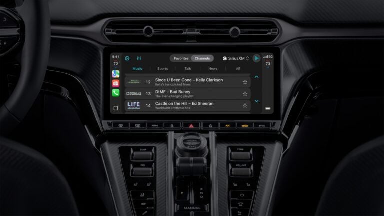 Mittelkonsole eines Autos mit großem Touchscreen-Display, das die SiriusXM-Musik-App zeigt. Darauf sind Musiktitel von Kelly Clarkson, Bad Bunny und Ed Sheeran zu sehen. Unter dem Bildschirm befinden sich Bedienelemente für Klima und Sitzheizung sowie ein Drehknopf mit der Aufschrift „Engine Start“. Die Oberfläche hat eine Carbon-Optik.