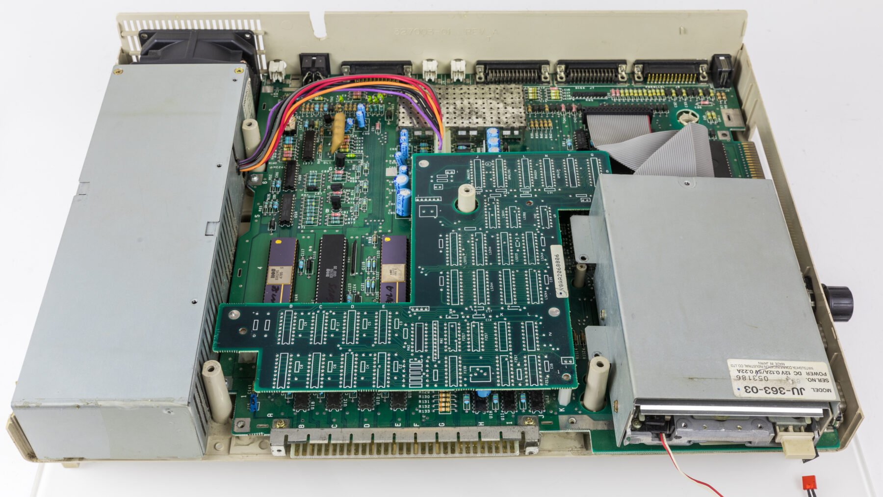 Geöffnete Elektronik mit sichtbarer Leiterplatte, mehreren integrierten Schaltkreisen, Kondensatoren, einem Lüfter links und einem Metallgehäuse rechts, das ein Laufwerk oder ein ähnliches Bauteil enthält. Mehrfarbige Kabel verbinden verschiedene Komponenten.