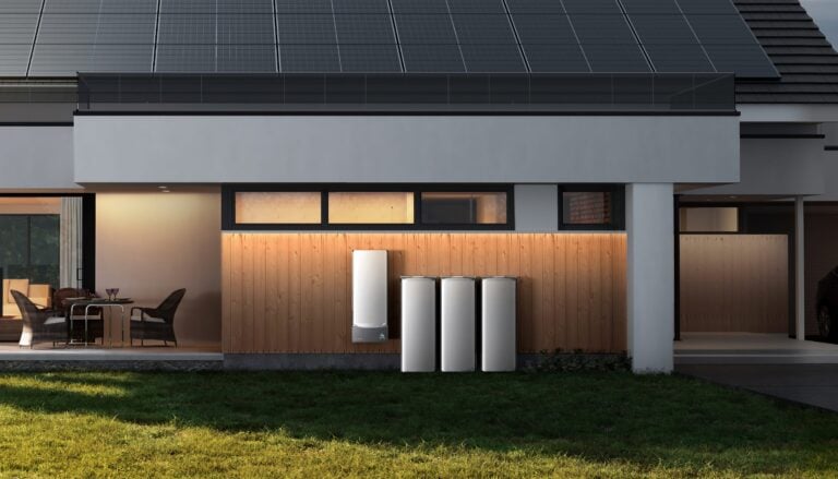 Moderne Hausfassade mit Holzverkleidung, drei weißen Energiespeicher- oder Batteriesystemen und einer Wandmontierten Einheit, darüber Fenster und ein Dach mit Solarpaneelen. Links eine überdachte Terrasse mit Sitzmöbeln, rechts eine Garage mit einem geparkten Auto. Vordergrund mit Rasen.