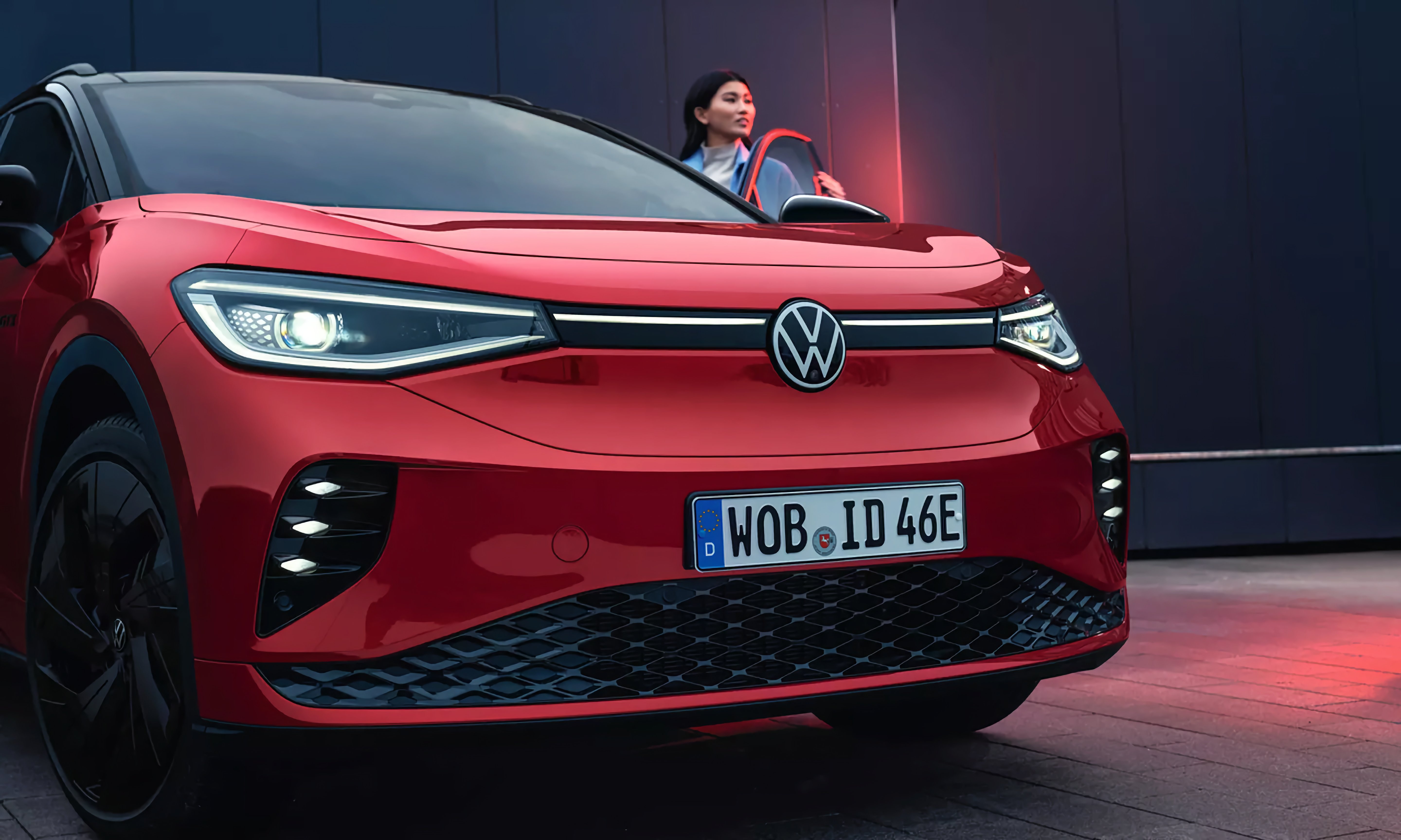 Roter Volkswagen ID.4 mit eingeschaltetem Scheinwerfer und schwarzem Kühlergrill, im Hintergrund eine Frau, die gerade die Fahrertür öffnet.