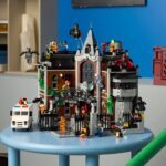 LEGO-Modell des Arkham Asylum mit mehreren Batman- und Joker-Figuren, auf einem blauen runden Tisch in einem Raum mit Regalen und Kisten im Hintergrund.