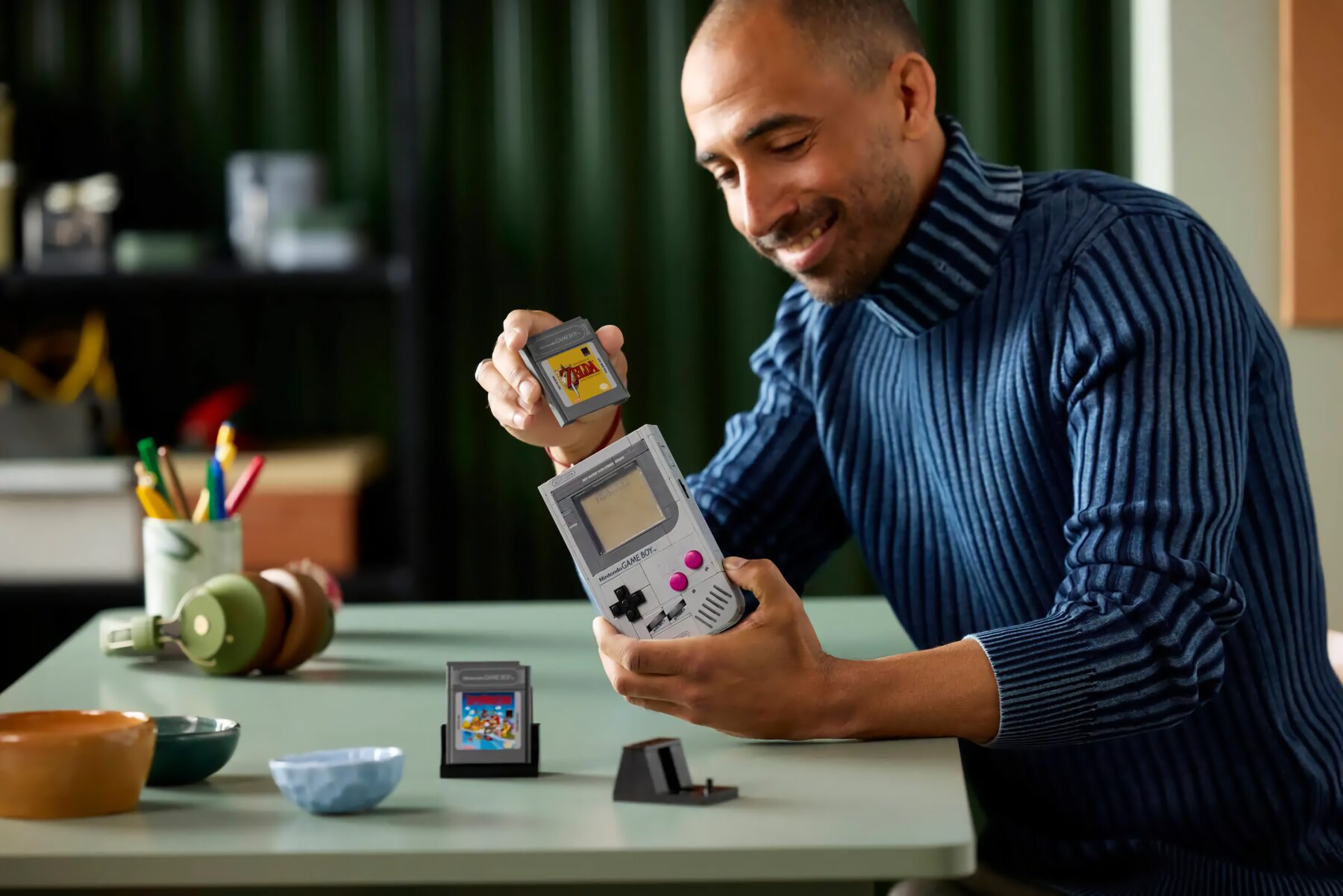 Mann in blauem Rollkragenpullover sitzt an einem Tisch und hält einen grauen Nintendo Game Boy sowie eine Spielkarte mit dem Spiel "The Legend of Zelda". Auf dem Tisch liegen weitere Spielkarten, bunte Stifte in einem Becher und grüne Kopfhörer.