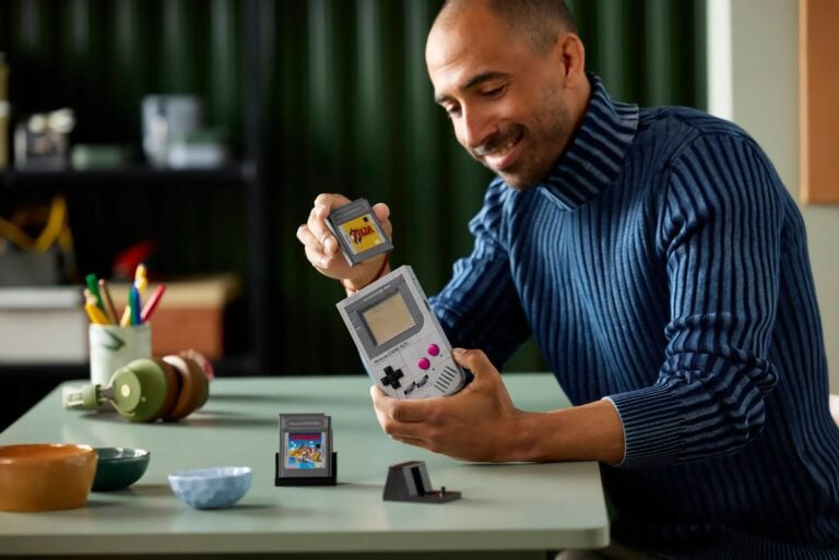 Mann in blauem Rollkragenpullover sitzt an einem Tisch und hält einen grauen Nintendo Game Boy sowie eine Spielkarte mit dem Spiel "The Legend of Zelda". Auf dem Tisch liegen weitere Spielkarten, bunte Stifte in einem Becher und grüne Kopfhörer.