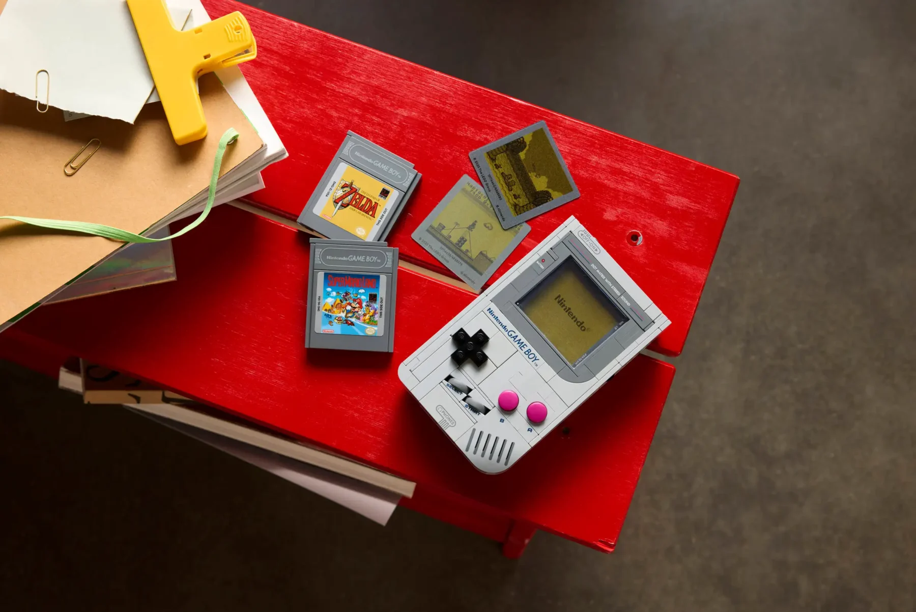 Ein grauer Nintendo Game Boy mit rosa Tasten liegt auf einem roten Tisch, daneben zwei Game Boy-Spiele mit den Titeln „The Legend of Zelda“ und „Super Mario Land“ sowie zwei kleine Spielkarten mit Pixelgrafiken. Links oben sind einige Papiere und ein gelber Clip zu sehen.