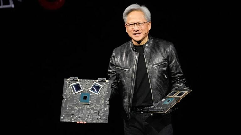 Ein Mann mit grauen Haaren und Brille trägt eine schwarze Lederjacke und hält zwei große Computerplatinen mit sichtbaren Prozessoren vor einem schwarzen Hintergrund.