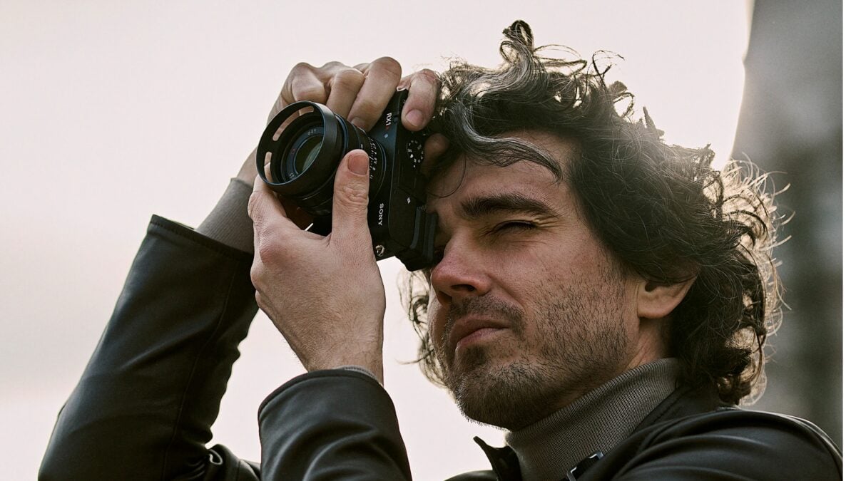 Ein Mann mit lockigem Haar und Bart hält eine Kamera nah an sein Gesicht und schaut durch den Sucher, während er fotografiert. Er trägt eine schwarze Lederjacke und einen grauen Rollkragenpullover.