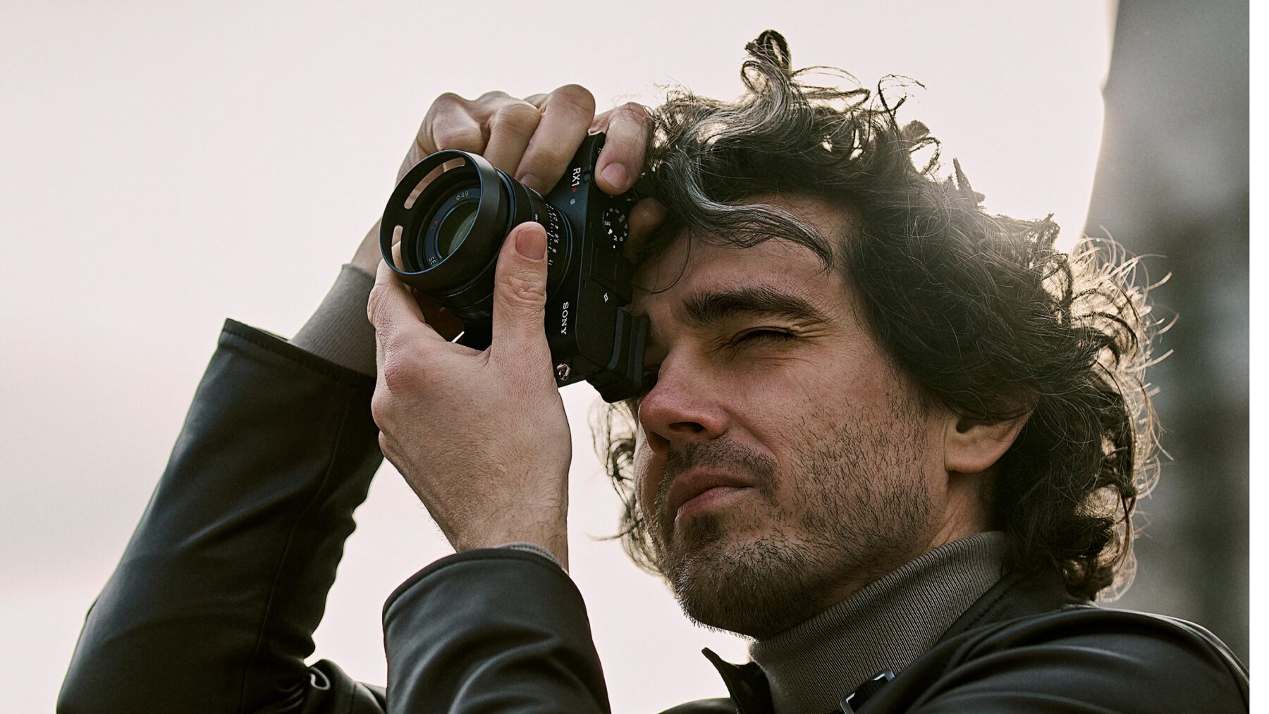 Ein Mann mit lockigem Haar und Bart hält eine Kamera nah an sein Gesicht und schaut durch den Sucher, während er fotografiert. Er trägt eine schwarze Lederjacke und einen grauen Rollkragenpullover.