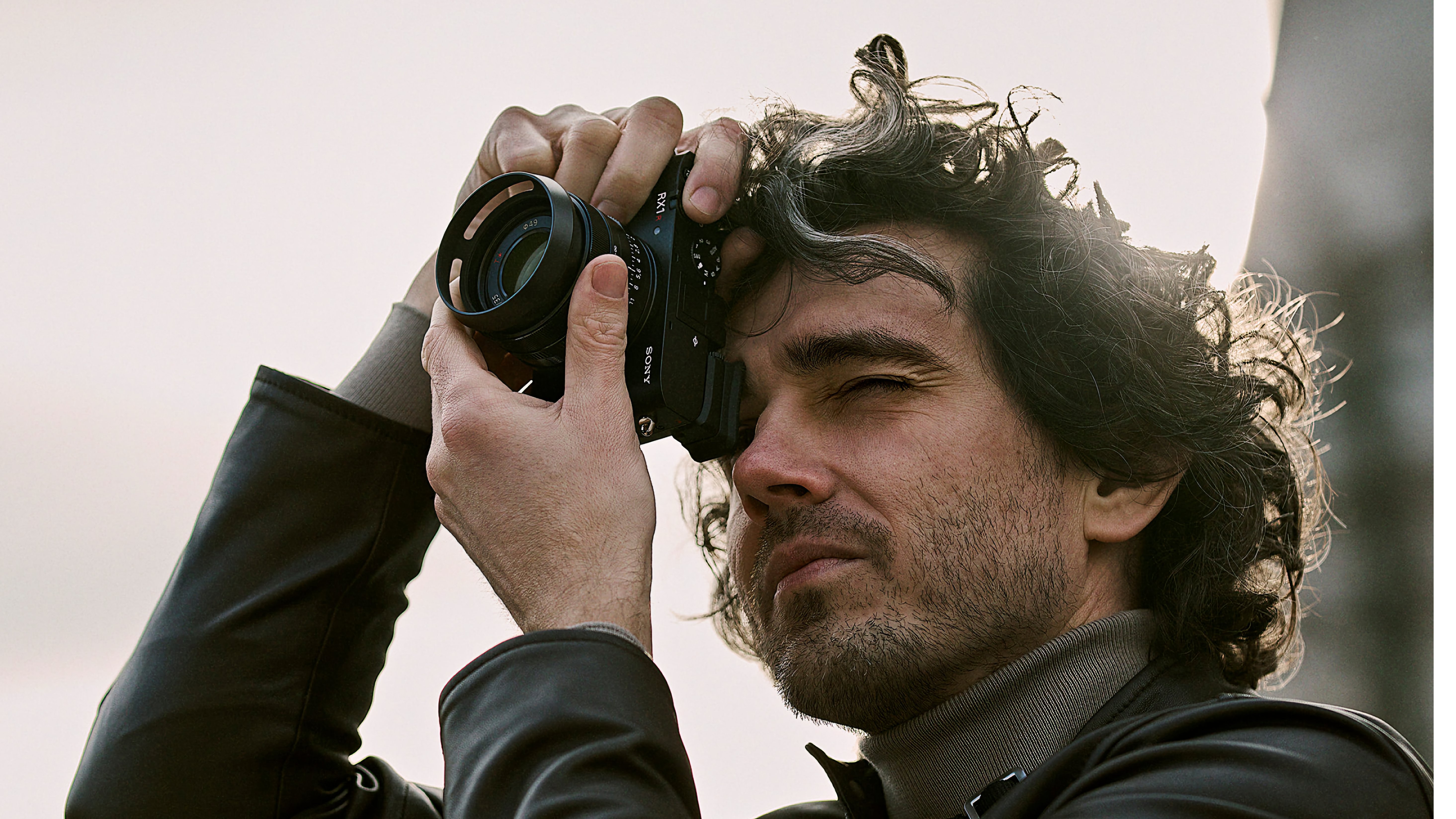 Ein Mann mit lockigem Haar und Bart hält eine Kamera nah an sein Gesicht und schaut durch den Sucher, während er fotografiert. Er trägt eine schwarze Lederjacke und einen grauen Rollkragenpullover.