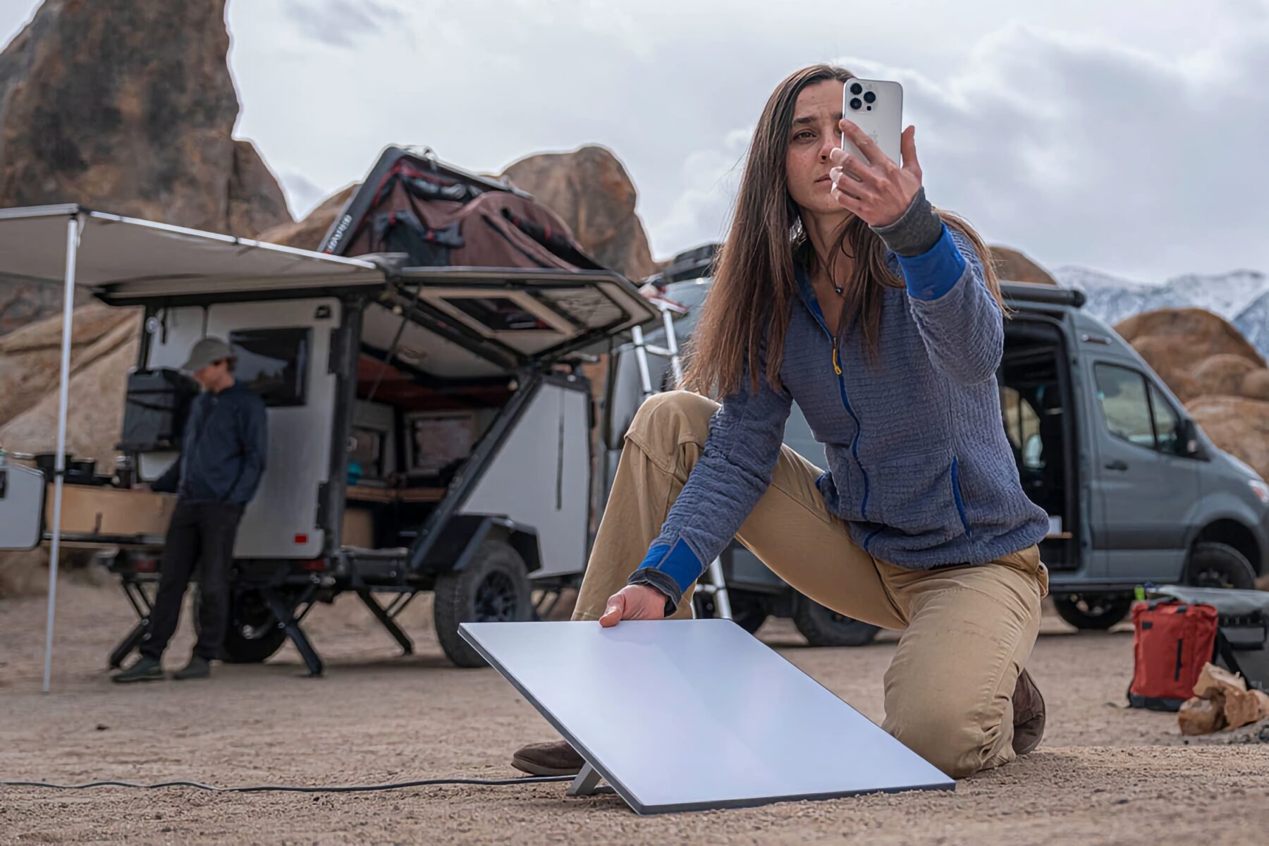 Frau kniet auf sandigem Boden und hält ein Smartphone, während sie ein flaches, rechteckiges Solarpanel aufstellt. Im Hintergrund stehen ein Camper-Van und ein Offroad-Anhänger mit geöffnetem Dachzelt, davor ein Mann mit Hut. Felsformationen und bewölkter Himmel sind sichtbar.