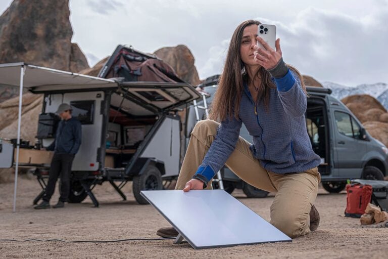 Frau kniet auf sandigem Boden und hält ein Smartphone, während sie ein flaches, rechteckiges Solarpanel aufstellt. Im Hintergrund stehen ein Camper-Van und ein Offroad-Anhänger mit geöffnetem Dachzelt, davor ein Mann mit Hut. Felsformationen und bewölkter Himmel sind sichtbar.