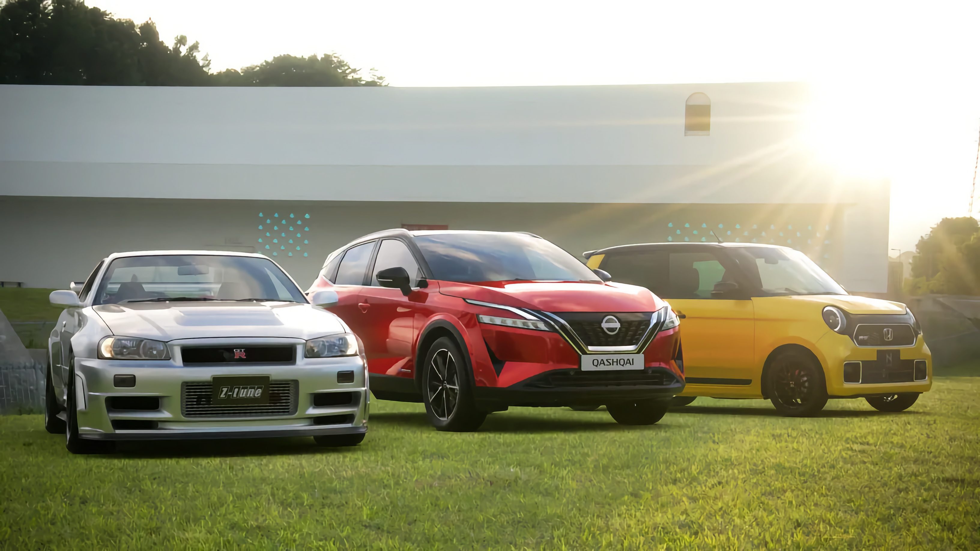 Drei Autos auf einer Wiese vor einem weißen Gebäude bei Sonnenuntergang: ein silberner Nissan Skyline GT-R, ein roter Nissan Qashqai und ein gelber Honda e.